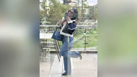 Stylish woman in blue jeans and a white fuzzy jacket taking a mirror selfie outdoors in a bright urban setting.