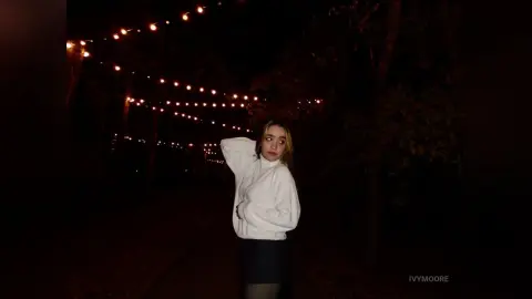 Young woman in a white sweater standing under glowing string lights in a dark, moody outdoor setting.