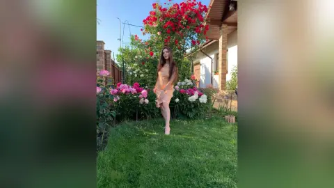Young woman in a peach summer dress standing in a lush garden filled with blooming pink and red roses.