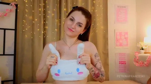 PetrinaFranceski wearing a white bunny-themed tube top, showing her tattoos and dark hair against a backdrop of warm fairy lights.