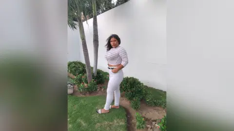 Young woman in a white long-sleeve top and white jeans posing in a sunny garden setting.