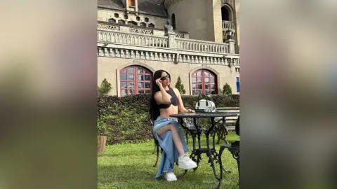 Stylish woman in a black crop top and denim skirt sitting at an outdoor cafe near a historic stone building.