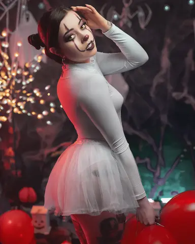 Woman in white bodysuit and tutu with clown makeup posing in a spooky Halloween setting with red balloons.