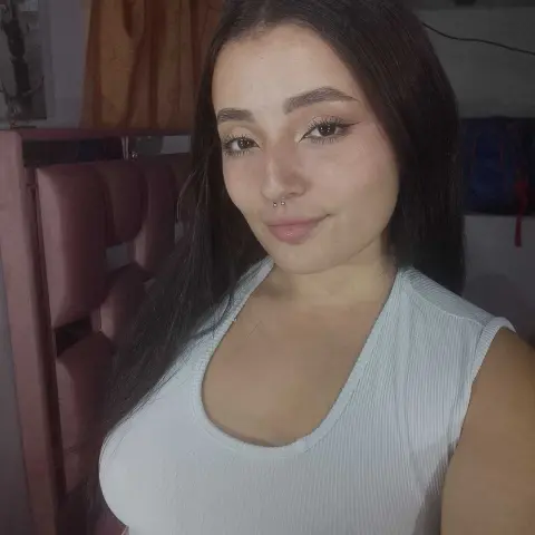 Young woman with long dark hair and nose ring wearing a white ribbed tank top in a cozy indoor setting.