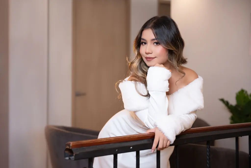 JessieOlivetti poses with her hands under her chin, wearing a white off-the-shoulder top and letting her wavy brown hair fall over her shoulders.