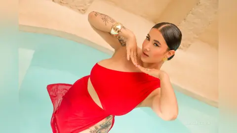 JuliahLewis poses in a red strapless dress against a light blue background, showing her tattoos and dark hair slicked back.