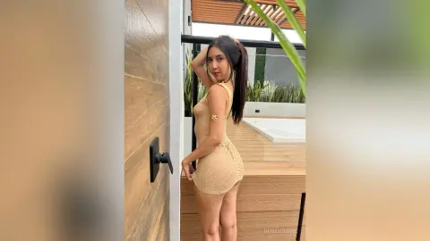 IrinaRussel poses in a tight, tan knit dress that hugs her curves, standing outdoors next to a wooden wall and a pool.