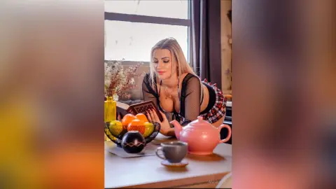 Blonde woman in a black mesh top reading a book by a table with fruit and a pink teapot in a cozy indoor setting.
