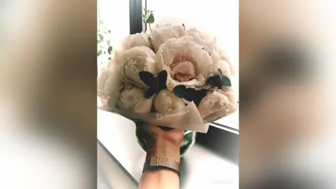 A close-up shot of a large bouquet of white peonies held in a hand against a bright window background.