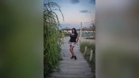 MissyJolie stands on a wooden boardwalk wearing a black tank top and white shorts against a backdrop of green bushes and a twilight sky.