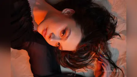 Young woman in a black dress lying on white bedding under warm, moody cinematic lighting.