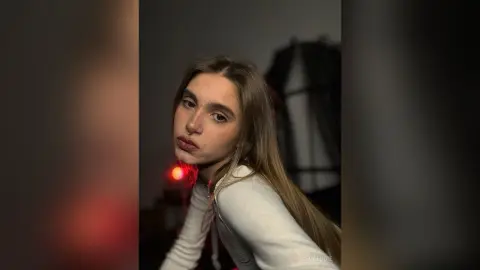 Young woman with long brown hair wearing a white long-sleeve top in a moody, dimly lit indoor setting.
