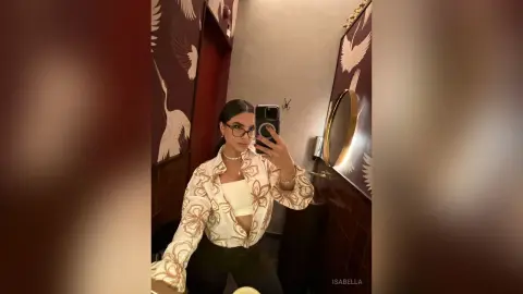Woman in white floral blouse and glasses taking a mirror selfie in a stylish room with bird wallpaper.