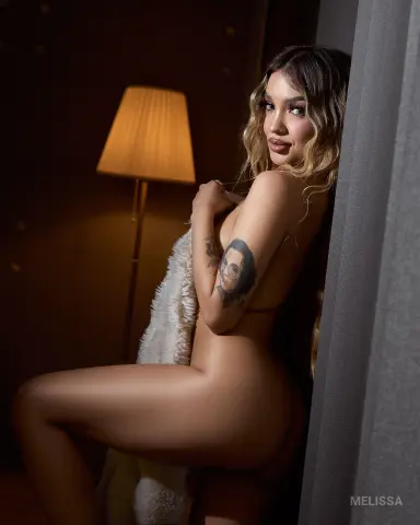 Young woman with long wavy hair and arm tattoo posing in a dimly lit room wrapped in a white faux fur blanket.