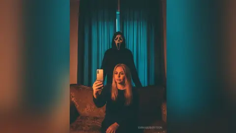 Spooky woman taking a selfie on a couch with a Ghostface killer looming behind her in a dark room.