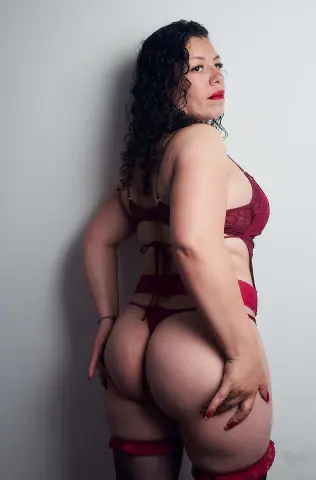 Curvy woman in red lace lingerie posing confidently against a plain white wall.