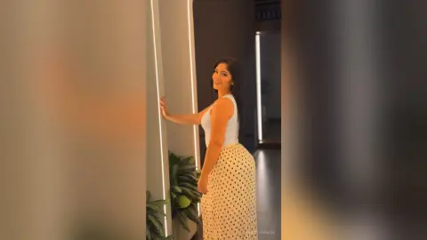 Smiling woman in a white sleeveless top and polka dot skirt posing in a modern dimly lit hallway.