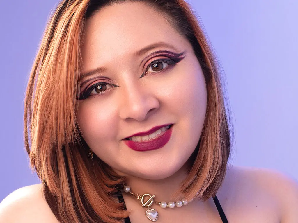 Smiling woman with reddish hair and bold winged eyeliner wearing a pearl choker against a purple background.
