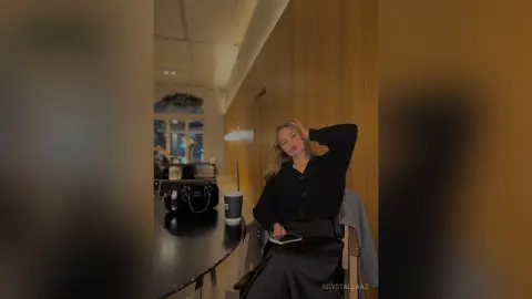 KrystalLaaz sits at a dark table in a dimly lit room, wearing a black long-sleeved shirt and looking toward the camera.