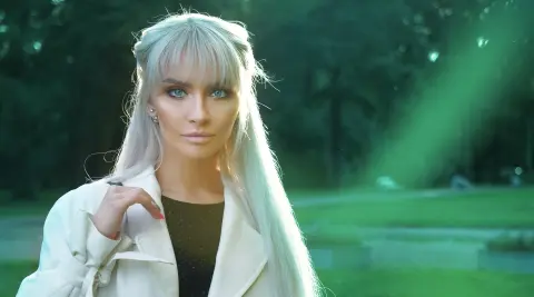 Blonde woman in a white blazer posing in a sunlit park with a serene expression.