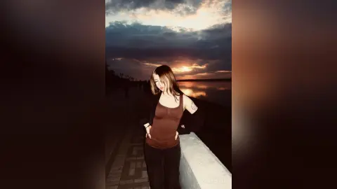 Young woman in a brown tank top standing on a pier under a dramatic sunset sky.