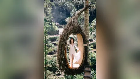 StacyHarland stands inside a large hanging nest or cocoon, wearing a long white dress against a lush tropical jungle background.