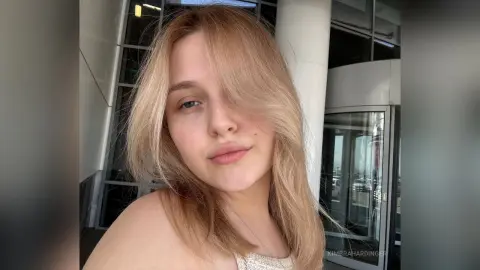 Blonde woman with long hair smiling in a modern glass building setting with natural lighting.