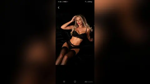 Blonde woman in black lace lingerie relaxing on dark fabric in a moody, intimate setting.