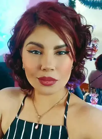 Queenamazon looks directly at the camera with red hair and red lipstick, wearing a black and white striped top and a gold necklace.