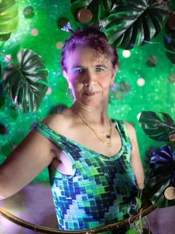 Smiling woman in a green patterned tank top posing against a tropical monstera leaf background with green lighting.