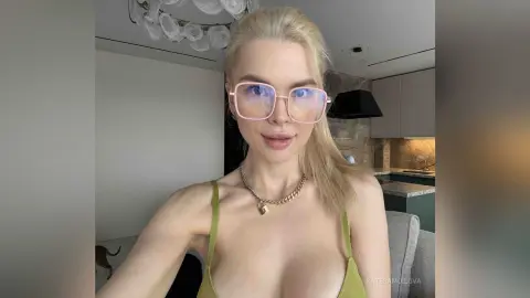 Blonde woman in a green tank top and pink glasses posing in a modern bright kitchen.