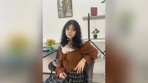 Young woman in a brown off-the-shoulder top and plaid skirt sitting in a bright, modern home office.