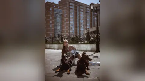 Stylish woman in a silver dress sitting on a city sidewalk with two brown dogs near urban buildings.