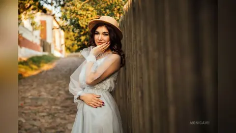 Dreamy portrait of a woman in a white sheer dress and tan hat leaning against a wooden fence in golden hour light.