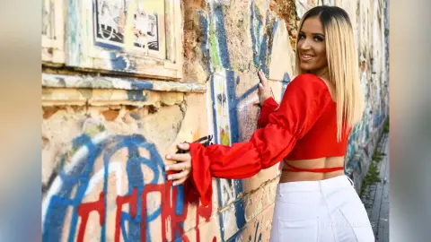TiffanyAddams poses against a graffiti-covered wall wearing a red long-sleeved crop top and white pants.