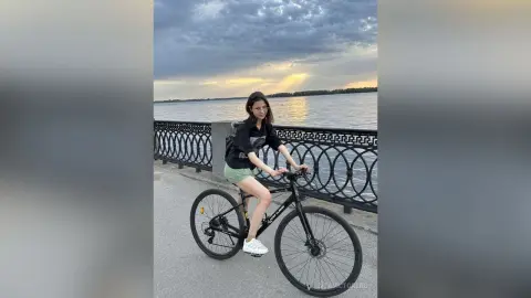 Young woman in casual black t-shirt and green shorts riding a bicycle by a lake under a cloudy sunset sky.
