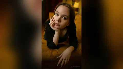Young woman in a black top leaning on a couch with a thoughtful, moody expression in warm indoor lighting.