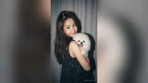 Smiling young woman in a black sequin dress holding a fluffy white Pomeranian dog in a studio setting.