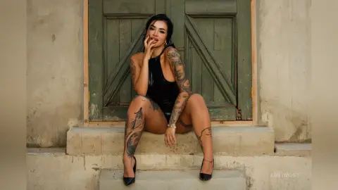 Tattooed woman in a black bodysuit sitting on stone steps in front of a rustic green door.