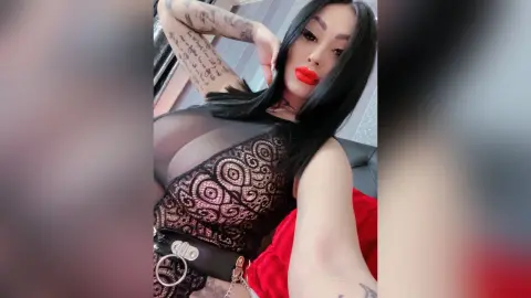 DeeaAnderson poses with long black hair and red lipstick, wearing a black lace bodysuit with a silver ringed belt.