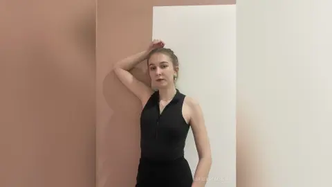 Young woman in a black sleeveless turtleneck top posing against a neutral wall in a minimalist setting.