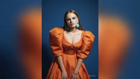 Fashion model in an elegant orange puff-sleeve gown posing against a blue studio background with bold blue eye makeup.