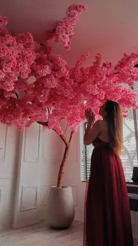 Woman in a long burgundy gown admiring a large pink artificial cherry blossom tree in a bright room.