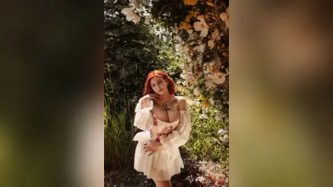 Redheaded woman in a white lace bohemian dress posing in a dreamy, sunlit garden filled with white flowers.