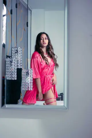 Young woman in a pink satin slip dress reflected in a mirror, creating a moody and intimate indoor aesthetic.