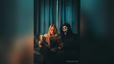 Blonde woman reading a Scream movie magazine next to Ghostface in a dark, moody living room setting.