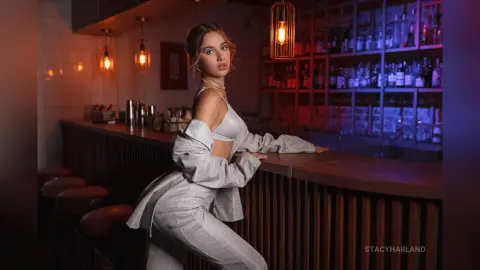 StacyHarland poses leaning against a dark wooden bar wearing a silver satin bra and matching trousers.