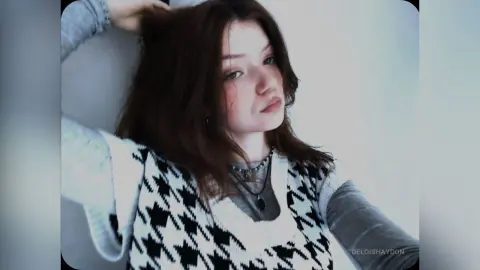 Young woman with brown hair wearing a patterned sweater vest and necklace in a soft, minimalist indoor setting.