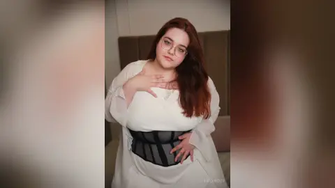 Smiling woman with glasses in a white blouse and black corset sitting in a cozy bedroom setting.