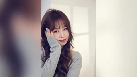 Smiling young woman with long wavy hair wearing a grey sweater in a bright, minimalist indoor setting.
